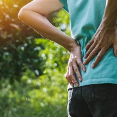 A person clutches their lower back as if in pain. They are outside with greenery in the background.